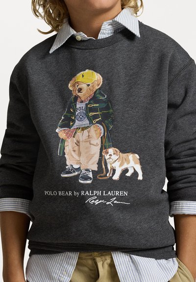 Siv sweatshirt z ilustracijo medveda v zeleni jakni in rumeni kapi, s kremnimi hlačami in majhnim psom ob njem.