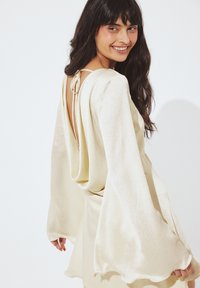 Obleka kremne barve z dolgimi, tekočimi rokavi, nazaj z zaveso in zavezovanjem na vratu, izdelana iz teksturirane, sijoče tkanine.