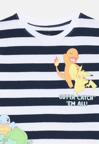 Jūras spēka un baltā striped t-kreklā ar zīmētiem varoņiem: Charmander, Pikachu un Squirtle; teksts lasa "Jānoķer visi!"