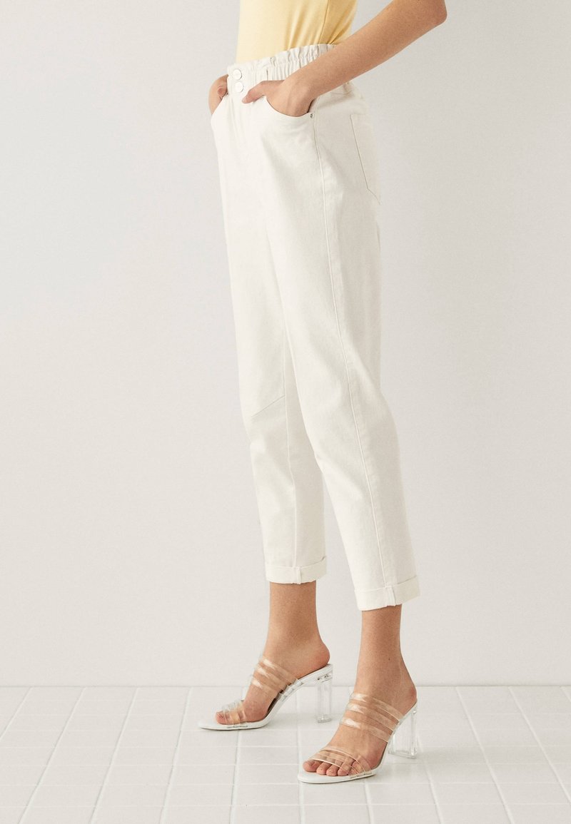 Une personne portant un pantalon taille haute blanc avec des ourlets repliés et des sandales à talons transparents, se tenant sur un sol carrelé blanc.