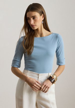 Γυναίκα με μακριά καστανά μαλλιά φοράει ανοιχτό μπλε τοπ με ανεστραμμένα μανίκια, λευκό παντελόνι ψηλής μέσης και πολλά βραχιόλια στον αριστερό καρπό.