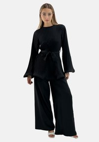 Haut noir plissé avec ceinture à nouer, associé à un pantalon noir à jambes larges. Le tissu est lisse, avec un design côtelé texturé.
