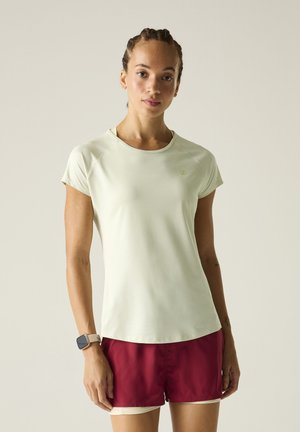 Joven mujer con el pelo trenzado lleva una camiseta de manga corta beige claro, pantalones cortos granate y un reloj inteligente, de pie frente a un fondo liso.