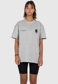 Graues, übergroßes T-Shirt aus Baumwolle mit einem schwarzen Logo auf der linken Brust und schwarzem Text auf der Vorderseite, kurzen Ärmeln und lockerer Passform.