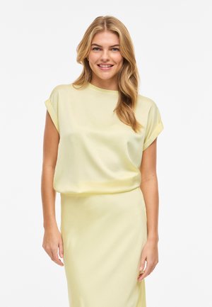 Lächelnde Frau mit langen blonden Haaren, die ein hellgelbes kurzärmeliges Oberteil und einen passenden Rock vor weißem Hintergrund trägt.