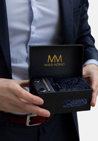 Coffret cadeau bleu marine contenant un mouchoir de poche à motifs, une pince à cravate métallique et des boutons de manchette ronds avec des accents bleus. Logo de la marque sur le coffret.