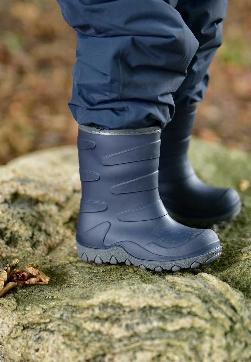 Bottes de pluie en caoutchouc gris avec une surface texturée, un design courbé et une semelle robuste à crampons. La hauteur atteint juste en dessous des genoux.