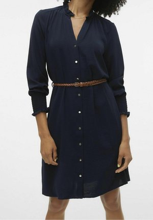 Kobieta ubrana w granatową sukienkę z przodu zapinaną na guziki, z marszczonymi mankietami i plecionym brązowym paskiem w talii, stojąca na jednolitym tle.