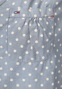 Tissu bleu clair à pois blancs avec une petite poche froncée ornée de détails cousus rouges, blancs et bleus dans le coin supérieur gauche.
