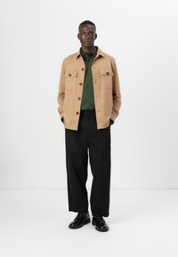 Veste en daim beige avec deux poches poitrine, chemise à boutons verte, pantalon noir ample et chaussures noires. Fond uni.