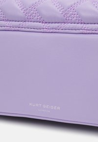 Purppura quiltattu nahkalaukku, jossa kontrastiva stitching, sileä tekstuuri ja kohokuvioitu merkin logo. Yksinkertainen, jäsennelty muotoilu.