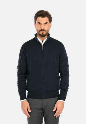 Maglione blu navy con zip e colletto alto, orlo e polsini a coste, tessuto texture, indossato sopra una camicia bianca, abbinato a pantaloni grigi.