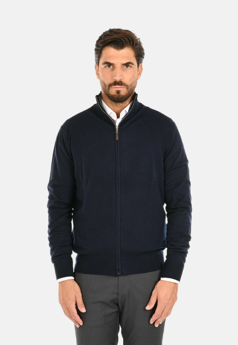Maglione blu navy con zip e colletto alto, orlo e polsini a coste, tessuto texture, indossato sopra una camicia bianca, abbinato a pantaloni grigi.