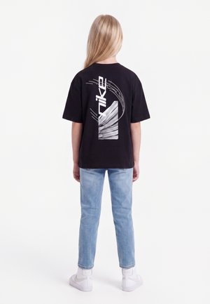 Enfant aux longs cheveux blonds, vu de dos, portant un t-shirt noir Nike, un jean bleu clair, des chaussettes blanches et des baskets blanches.