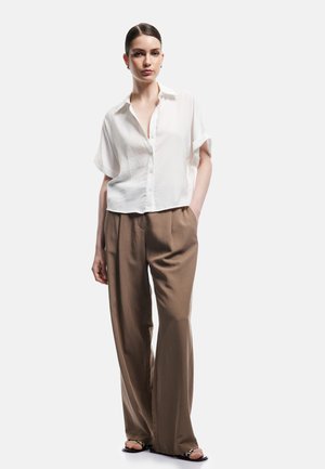Kobieta w luźnej białej koszuli z krótkim rękawem zapinanej na guziki, brązowych szerokich spodniach i czarnych sandałach, stoi z jedną ręką w kieszeni.