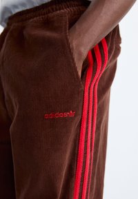 Brown corduroy pants with red three-stripe accents and Adidas logo. Features elastic waistband and side pockets. Textured, loose fit.