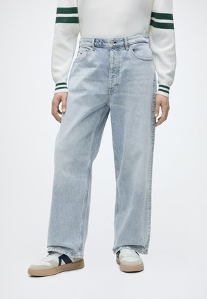 Osoba nosząca jasnoniebieskie szerokie jeansy, białe trampki z beżowymi podeszwami i czarnymi akcentami oraz białą koszulę z długim rękawem w zielone paski.