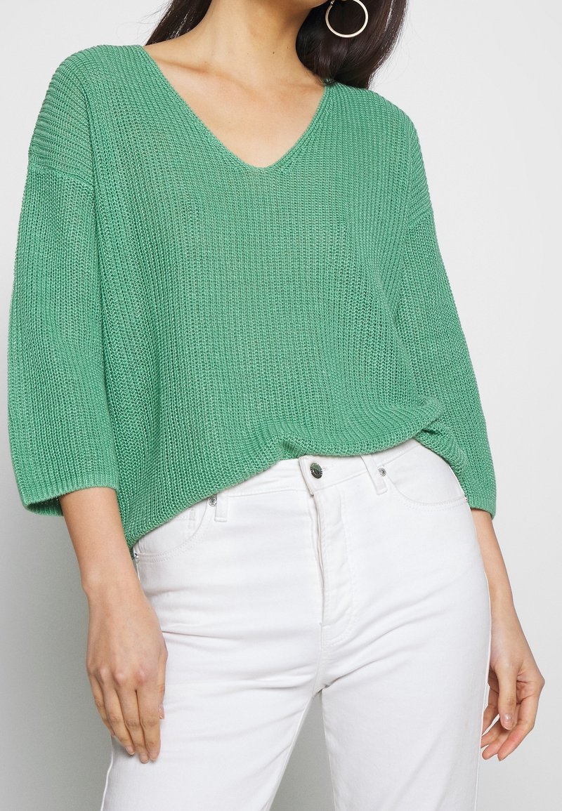 Femme portant un pull en maille vert ample avec des manches trois-quarts et un pantalon blanc taille haute, accessoirisée avec de grandes boucles d'oreilles en anneau.