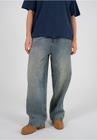 Vaqueros de pierna ancha en azul claro desgastados, combinados con una camiseta oversize azul marino y calzado marrón con suela texturizada.