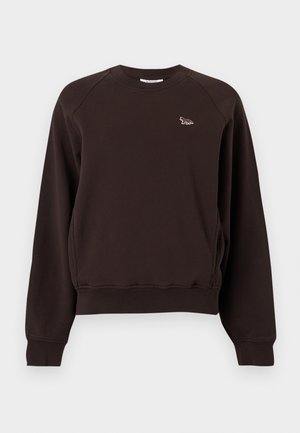 Kobieta w ciemnobrązowej bluzie, beżowych szortach i beżowych sandałach z kokardkami, stoi z jedną ręką w kieszeni na jednolitym białym tle.