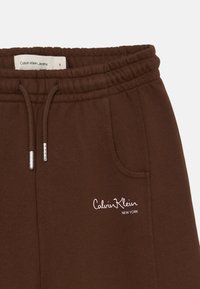 Calças de brim castanhas com cintura elástica, cordão, bolsos laterais e o logotipo "Calvin Klein" bordado na parte da frente.