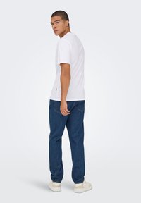 White short-sleeve t-shirt, relaxed fit, paired with dark blue denim pants. Notable detail: small logo accent on the shirt's side.
