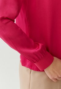 Blusa de satén fucsia con mangas largas, puños gathered y textura suave, combinada con pantalones beige claros.