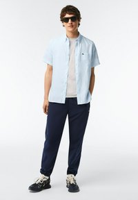 Camicia azzurra a maniche corte con bottoni, t-shirt grigia sotto, pantaloni da jogging blu scuro con polsini elastici e scarpe nere con lacci bianchi e dettagli bianchi.