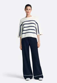 Fehér, bolyhos pulóver sötétkék csíkokkal, háromnegyedes ujjú, sötétkék széles szárú nadrággal, fehér díszítéssel. A lábbeli texturált mintázatú.