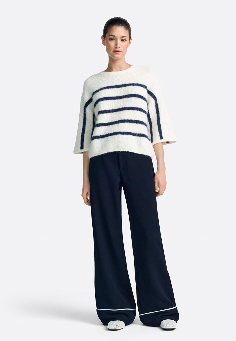 Fehér, bolyhos pulóver sötétkék csíkokkal, háromnegyedes ujjú, sötétkék széles szárú nadrággal, fehér díszítéssel. A lábbeli texturált mintázatú.