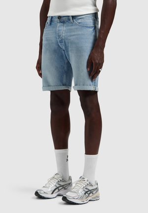 Man draagt lichtblauwe opgerolde spijkershorts, witte sportsokken, zilveren hardloopschoenen en een wit mouwloos shirt, staand op een effen achtergrond.