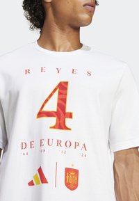 Hvid bomulds T-shirt med kraftige røde og gule grafikker. Designet inkluderer nummeret "4" med et bølgemønster, tekst og brandinglogo.