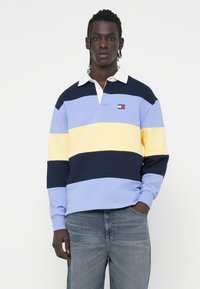 Maglietta tipo polo a strisce blu, gialle e navy con colletto bianco. Presenta un piccolo logo sul petto. Realizzata in tessuto di cotone.