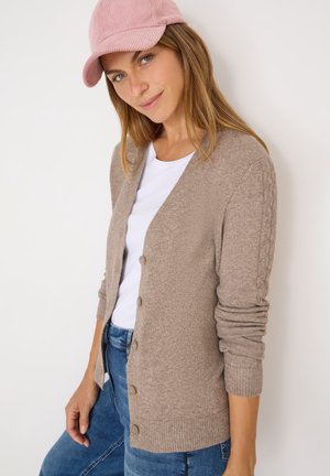 Helles beige Cardigan mit strukturierter Strickware, zentralem Knopfverschluss und gerippten Bündchen. Über einem weißen Hemd und blauen Jeans getragen.