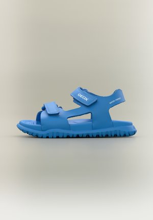 Sandalia azul para niños con correas ajustables, suela resistente, etiquetada con "GEOX" y "WATER FRIENDLY" en los lados, sobre un fondo neutro.