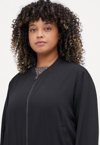 Black bomber jacket with a zippered front, relaxed fit, and side pockets; features a textured fabric and a round collar.