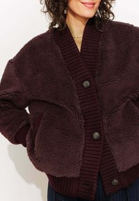 Cardigan en polaire surdimensionné bordeaux avec des poignets et un col côtelés, présentant de grandes poches avant et deux boutons marron. Tissu doux et texturé.