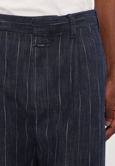 Donkerdenim broek met verticale witte lijntjes. Kenmerkt zich door een tailleband met lussen en een klein "CLOSED" label nabij de taille.