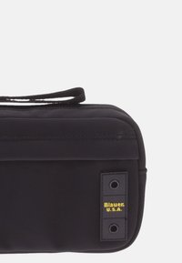 Bolsa de tela negra con cierre de cremallera, que presenta una etiqueta con texto en amarillo. Textura suave, forma rectangular y un pequeño asa para llevar.