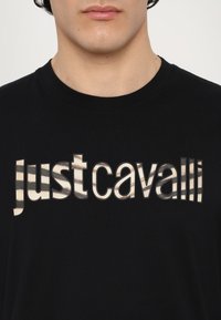 La maglietta nera presenta la scritta "justcavalli" in un print metallizzato bold e a righe. Il tessuto appare liscio e simile al cotone.