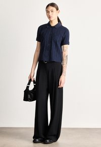 Tumši zila īsroku krekls ar teksturētu rakstu un pogām, kopā ar augsti piegulošiem melniem platajiem biksēm un nelielu melnu rokassomiņu.