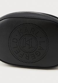 Fekete bőr kozmetikai táska perforált logó dizájnnal, ovális forma, cipzáros záródás, sima felülettel. Ezüst cipzárral díszítve.