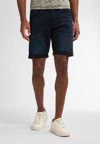 Tummansinisiä farkkushortseja, joissa on käännetty saumateko, sisältäen klassisen viisi-taskuisen muotoilun. Yhdistettynä valkoisiin lenkkareihin rennon ilmeen vuoksi.