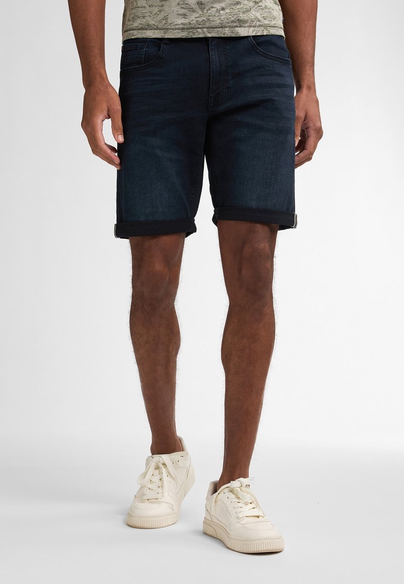 Tummansinisiä farkkushortseja, joissa on käännetty saumateko, sisältäen klassisen viisi-taskuisen muotoilun. Yhdistettynä valkoisiin lenkkareihin rennon ilmeen vuoksi.