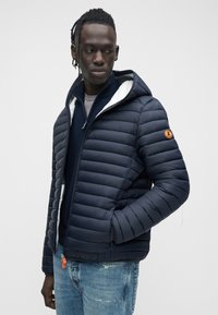 Tengerészkék puffertalpú dzseki kapucnival, cipzáras záródással és bordázott mandzsettákkal. Fehér béléssel és logópálcával az ujjon. Farmerrel viselve.