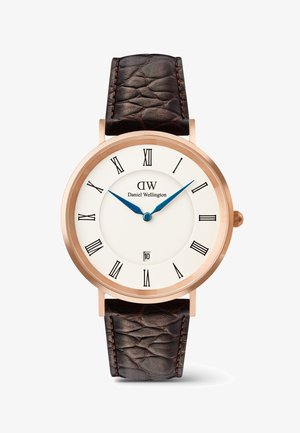 Okrągły zegarek w kolorze różowego złota z białą tarczą, czarnymi rzymskimi cyframi, niebieskimi wskazówkami, okienkiem daty na godzinie 6 oraz ciemnobrązowym, teksturowanym paskiem ze skóry.