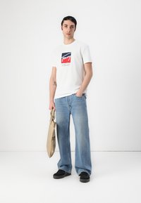Biela bavlnená tričko s retro logom, skombinované s svetlomodrými širokými džínsami a čiernymi teniskami. Model drží hnedú tašku.