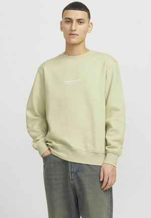 Jasnozielony sweatshirt z okrągłym dekoltem i ściągaczami przy rękawach. Na przodzie nadrukowany tekst "Originals Studio". Noszony z luźnymi dżinsami.