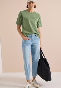 Femme portant un t-shirt vert à manches courtes, un jean bleu clair, des baskets blanches, une casquette beige, tenant un grand sac cabas noir d'une main.