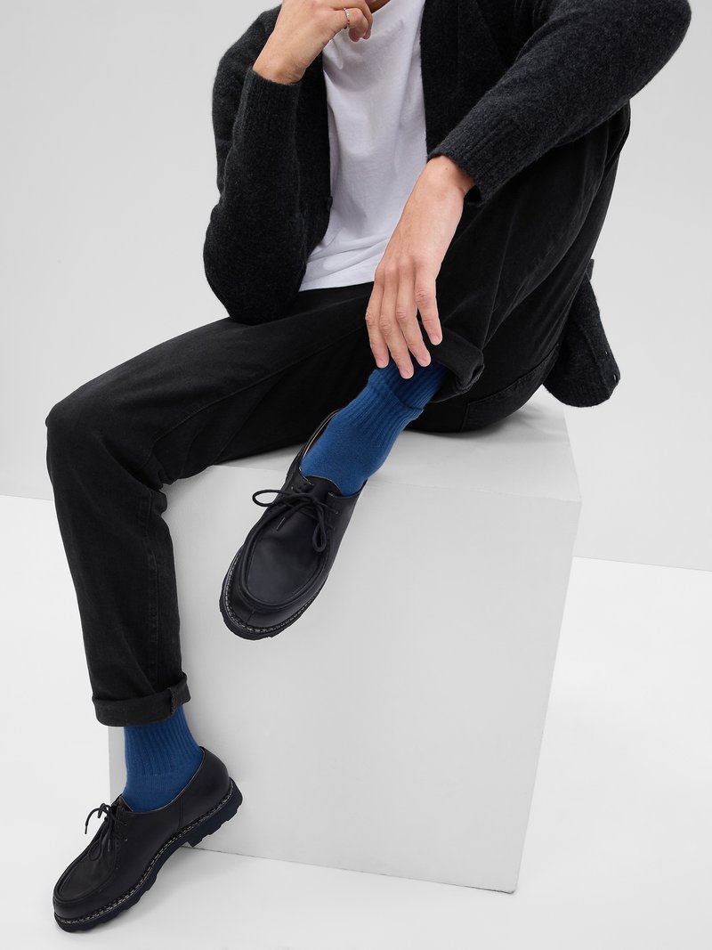 Des chaussures en cuir noir à lacets avec une surface texturée. Modèle portant des chaussettes côtelées bleues, un jean noir et un cardigan sombre sur une chemise blanche.